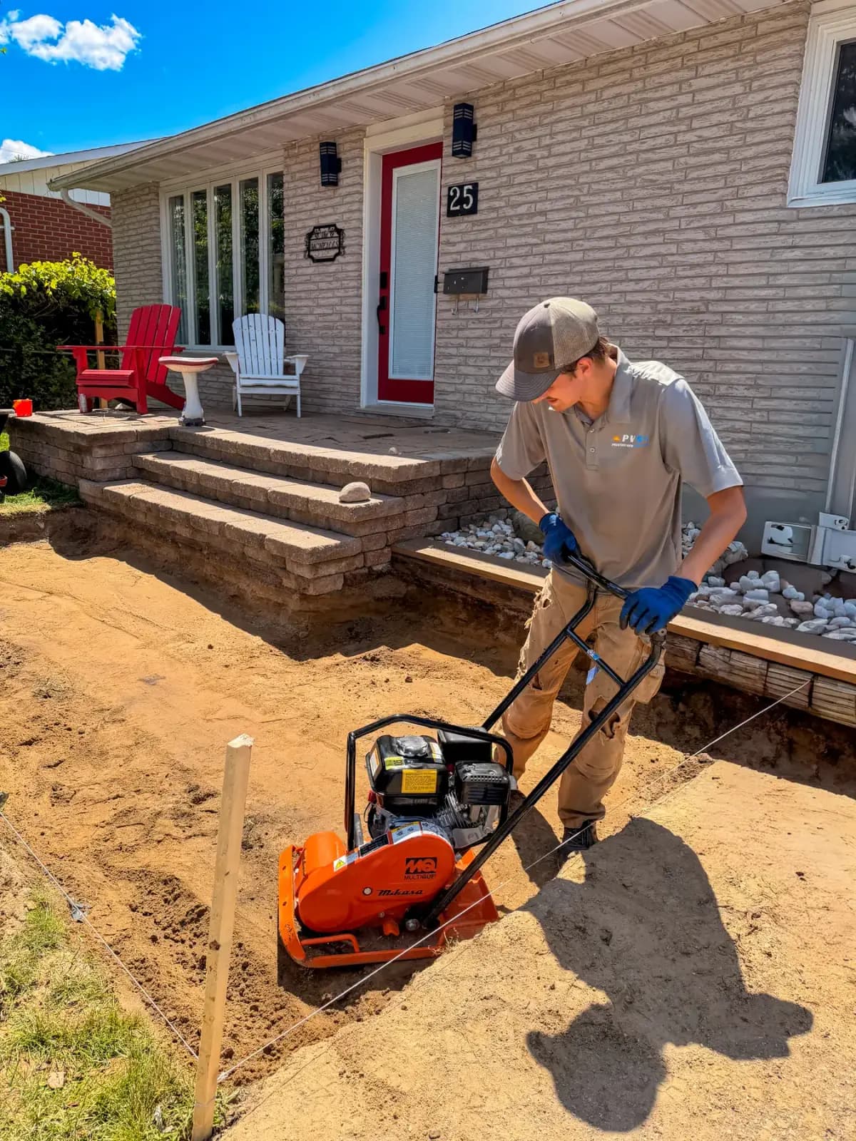 Crew member compacting the gravel base beside the stone front steps before laying pavers