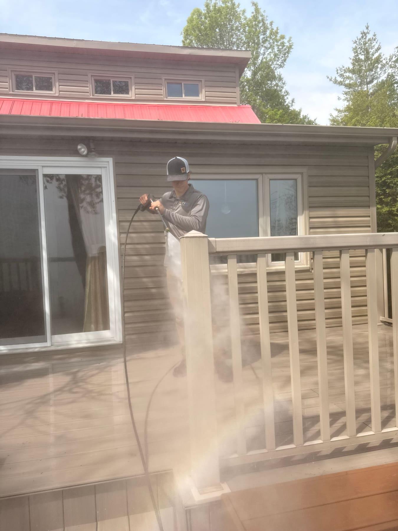 PVS technician in branded gear pressure-washing the second-storey siding of a home with a red metal roof — visible water mist captures the action mid-spray.